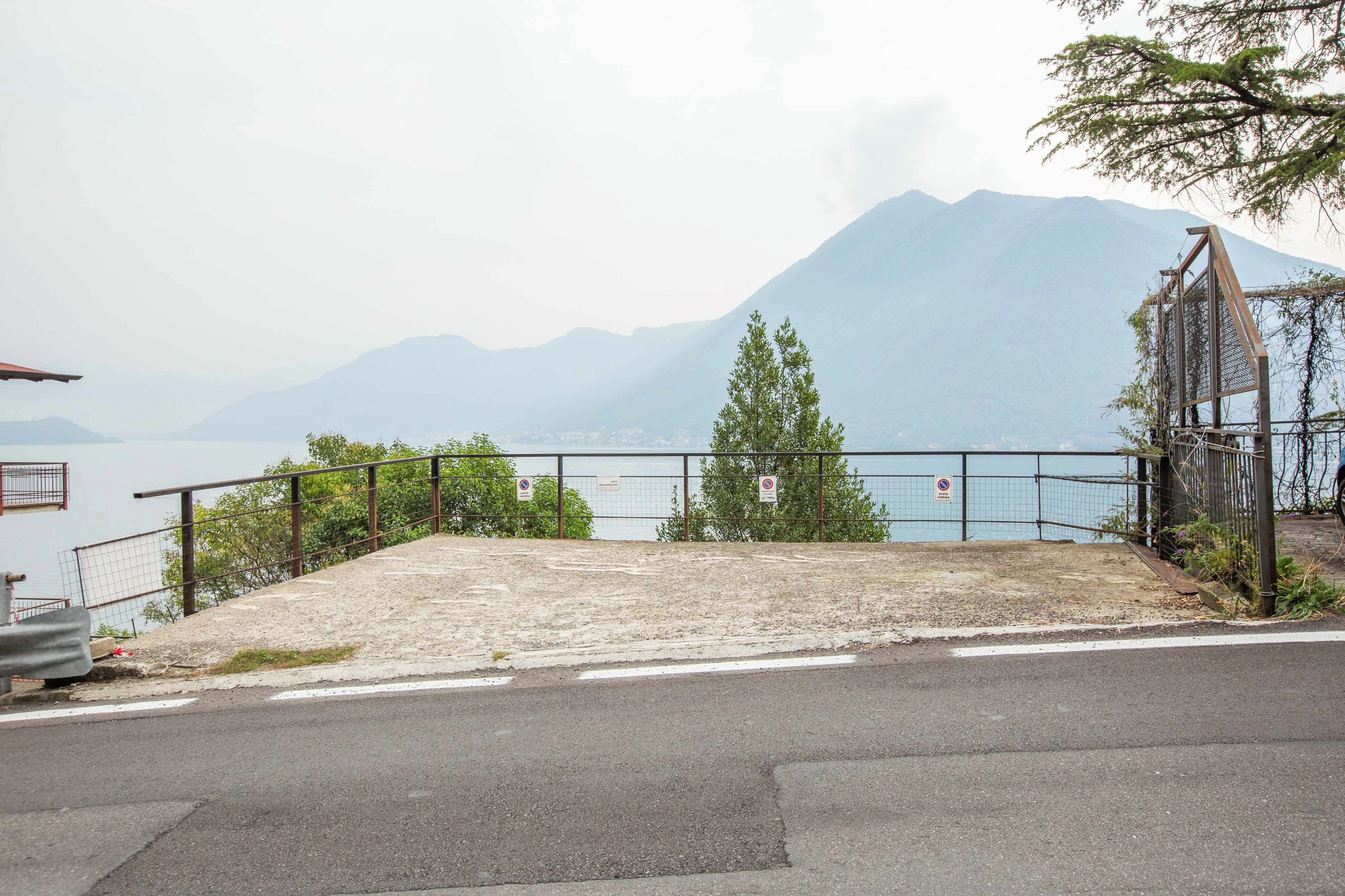 A paved area with a metal fence, a small tree, and a view of a lake with mountains in the distance.