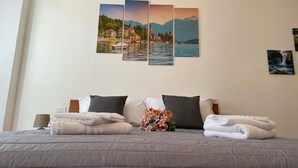 A neatly made bed with folded towels and a bouquet of flowers, a lamp, and three paintings on the wall depicting scenic landscapes. - Dream Holiday  (Argegno)
