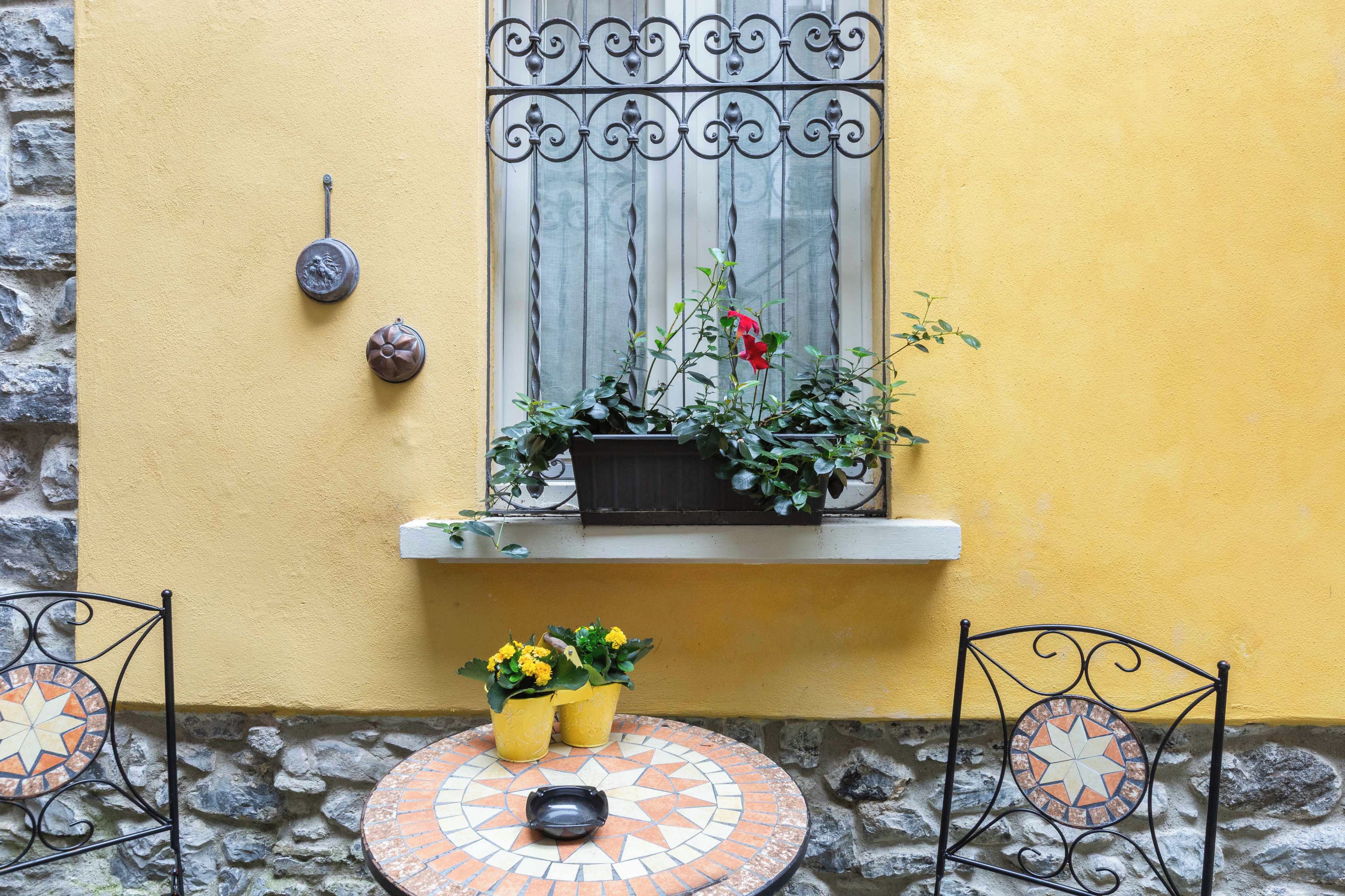 A yellow wall with a window, a stone wall, a metal gate, and a tiled table with a flower pot.