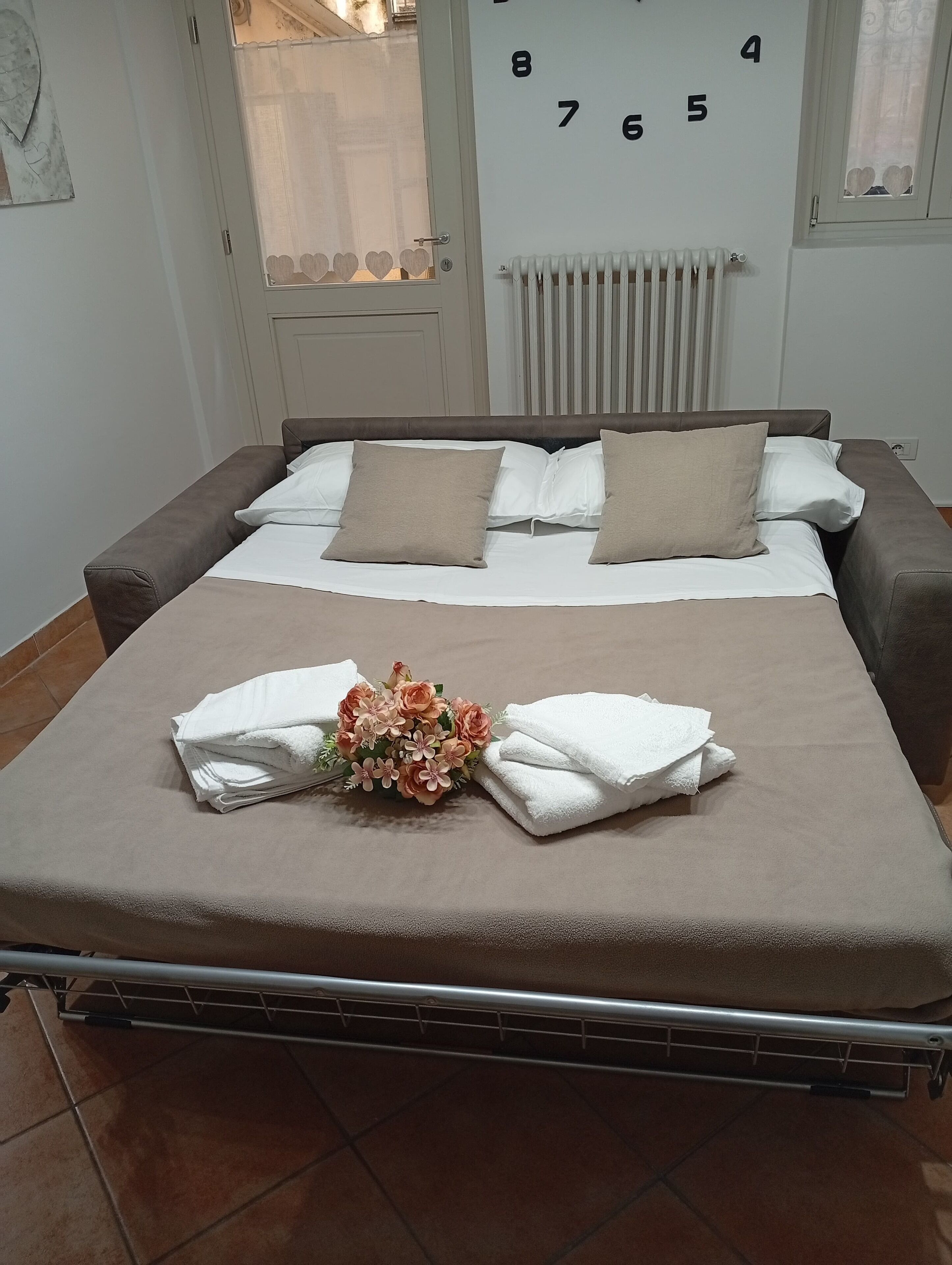 A neatly made bed with a floral arrangement and folded towels, a radiator, and a wall with numbers.