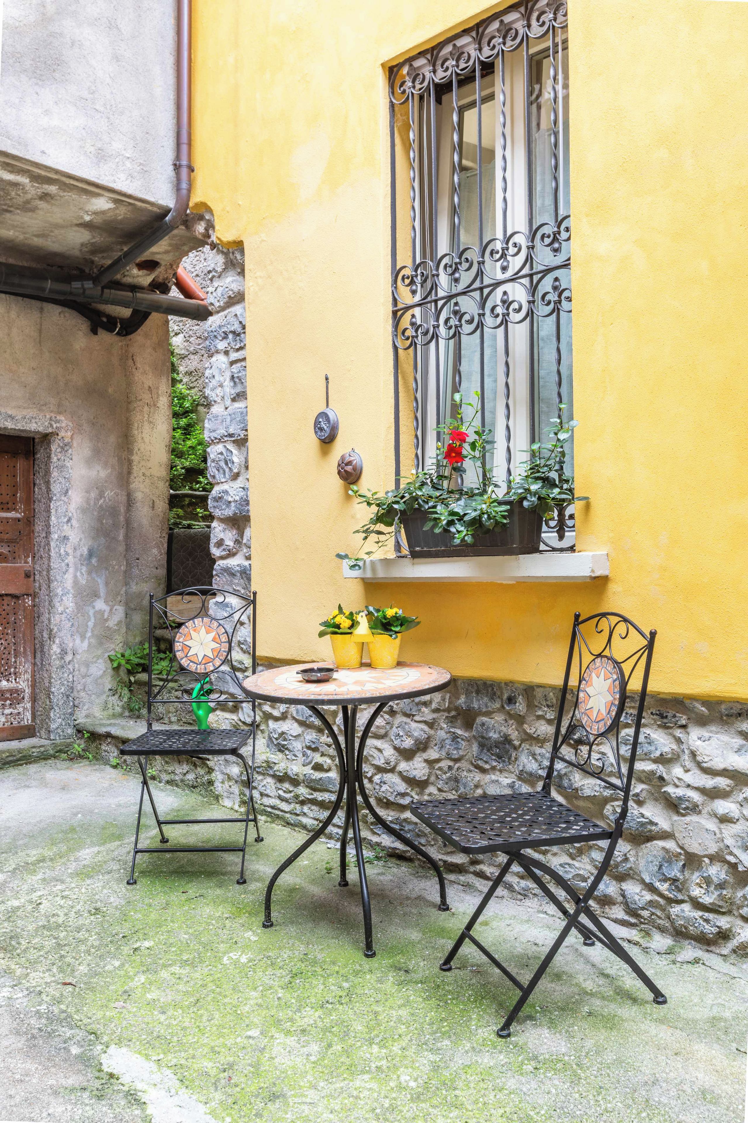 An outdoor seating area with a round table and two chairs, a window with decorative iron bars, and potted plants.