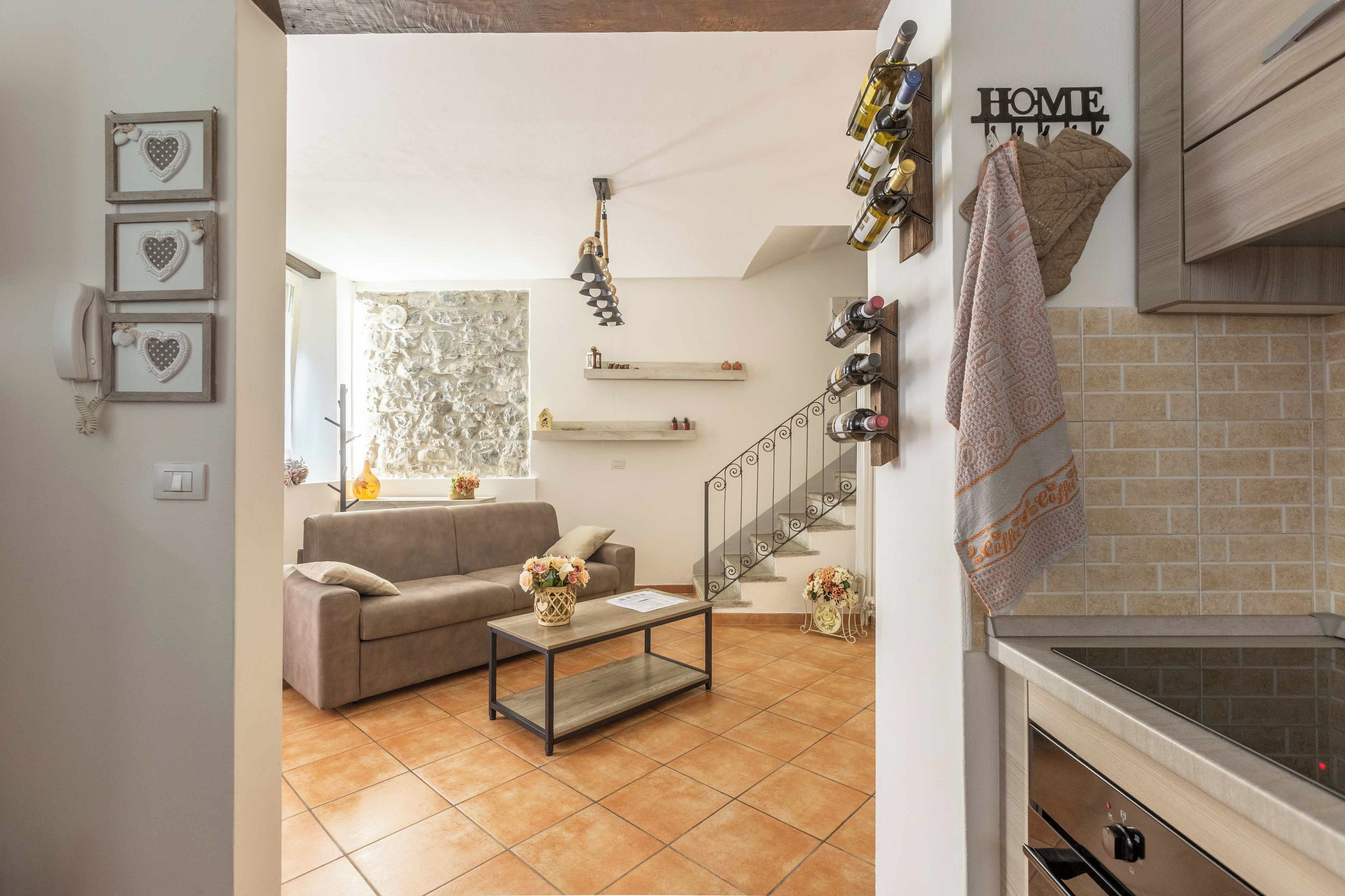 A modern living room with a sofa, coffee table, and decorative items. There is a staircase leading to another level, and a kitchen area with a stove.