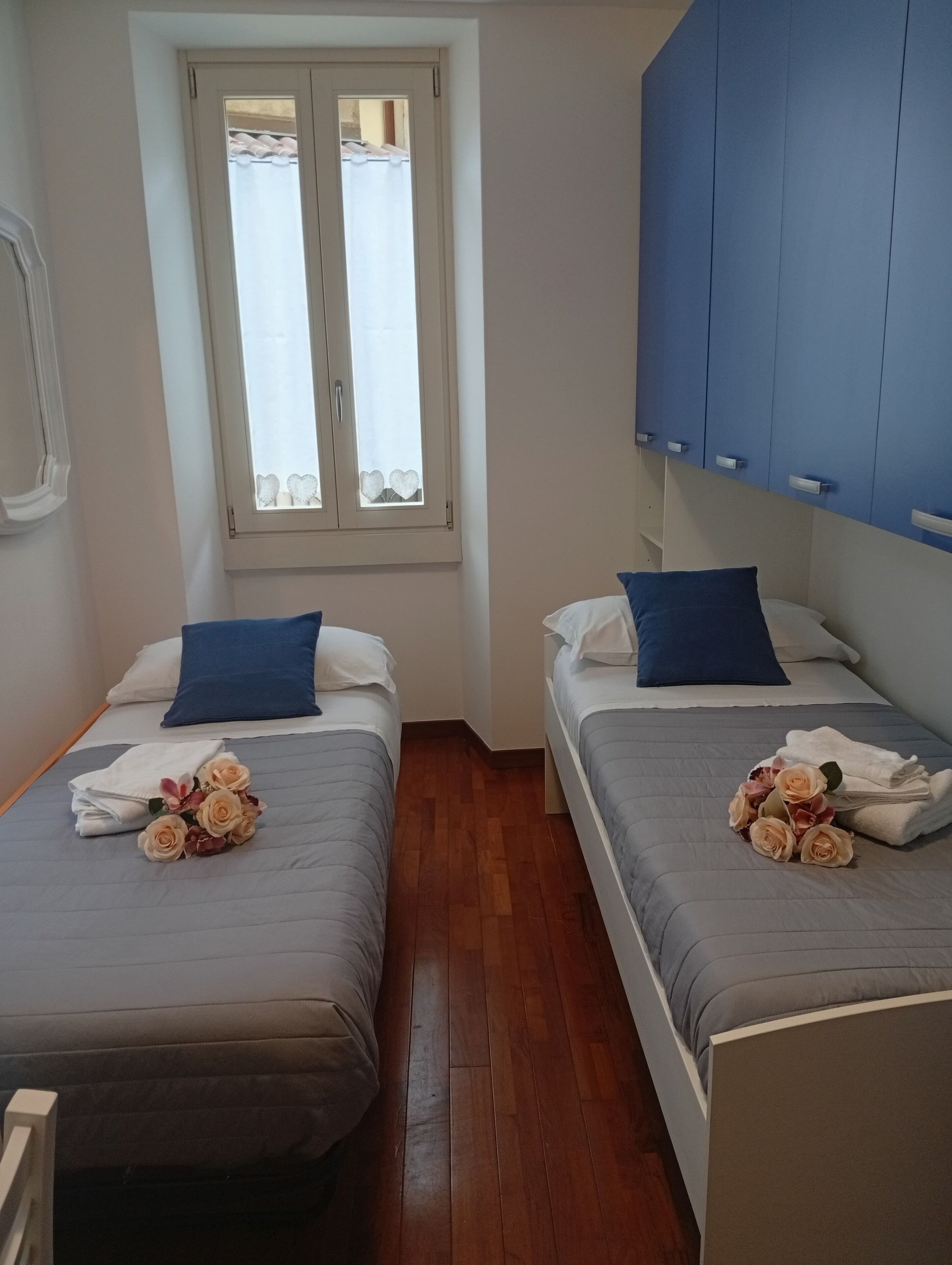A room with two beds, each with a blue pillow and a bouquet of flowers. There is a window with a curtain, and a blue wardrobe against the wall.