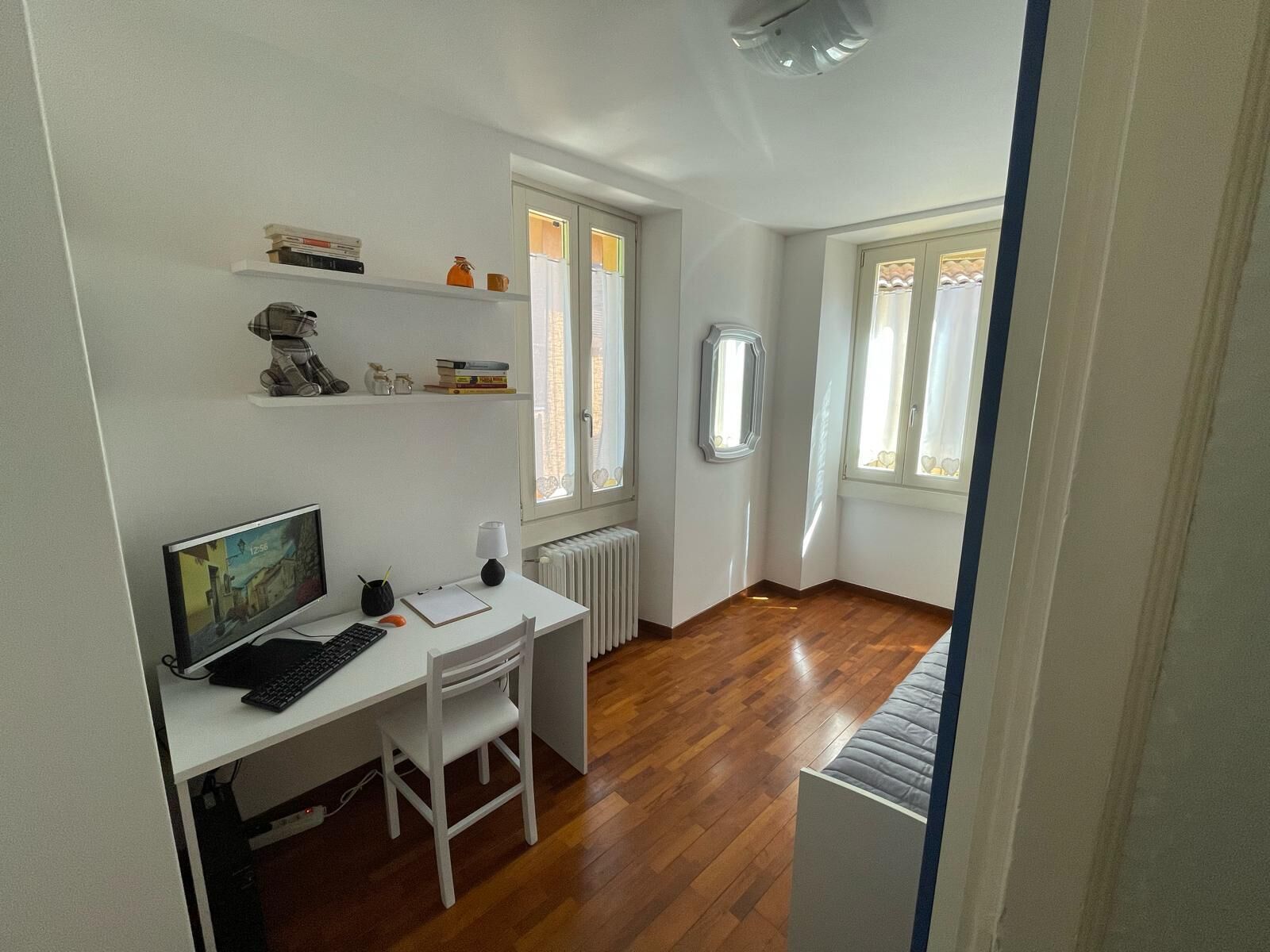 A room with a white desk, computer, and chair, a radiator, and two windows with curtains.