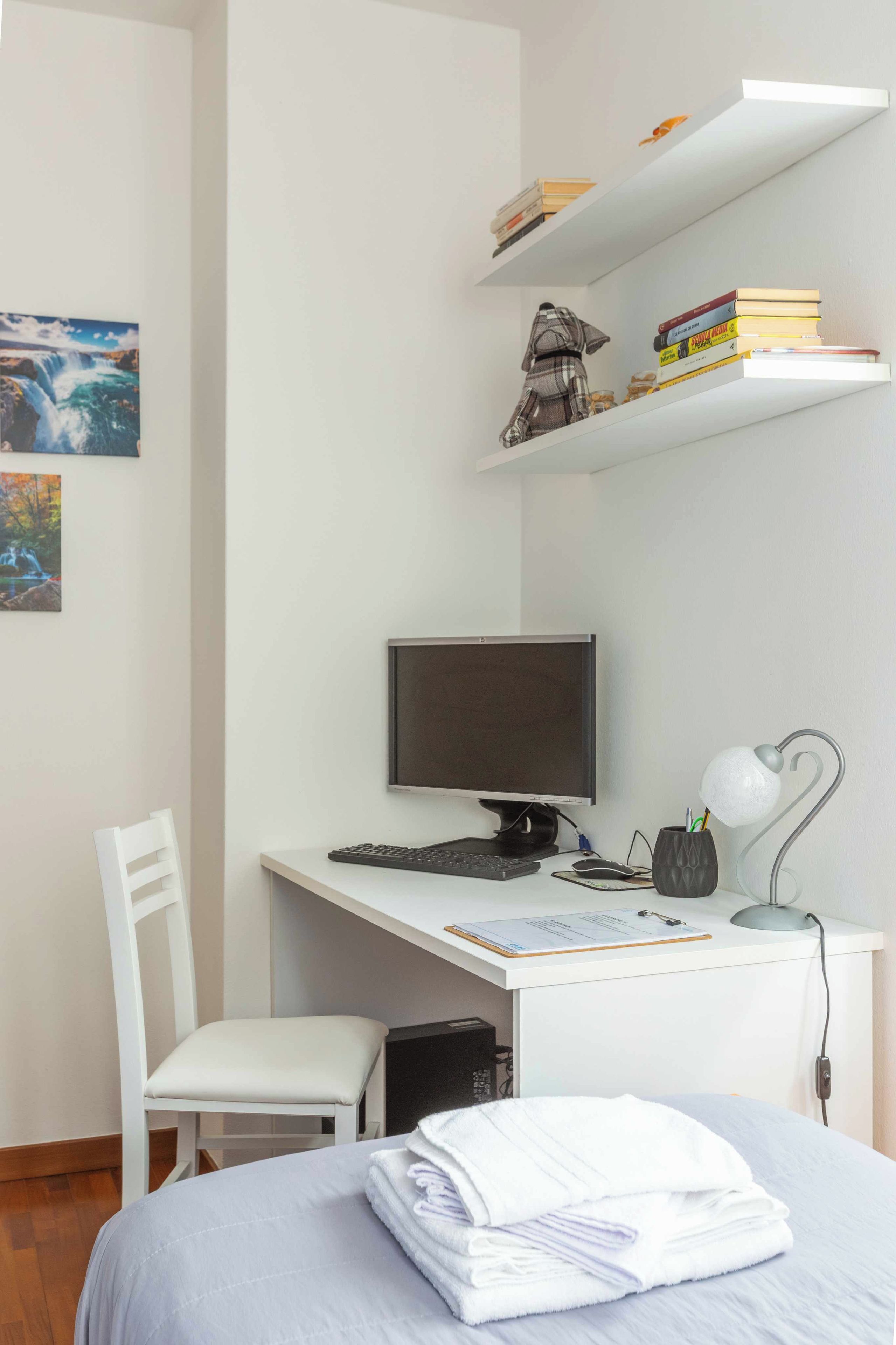 Un dormitorio con cama, escritorio con computadora, silla, estante con libros y un cuadro en la pared.
