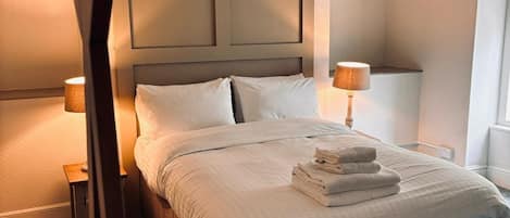 A neatly made bed with white linens and a stack of folded towels, a wooden headboard, two bedside lamps, and a carpeted floor.