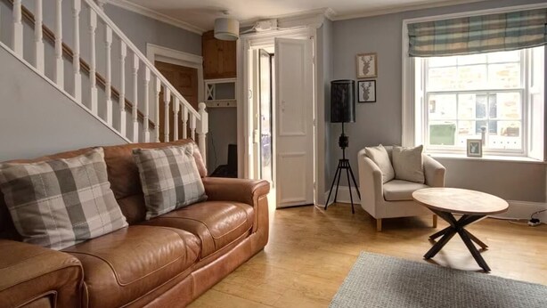 A living room with a brown leather sofa, a white armchair, a round wooden coffee table, a tripod, and a window with a view of buildings.