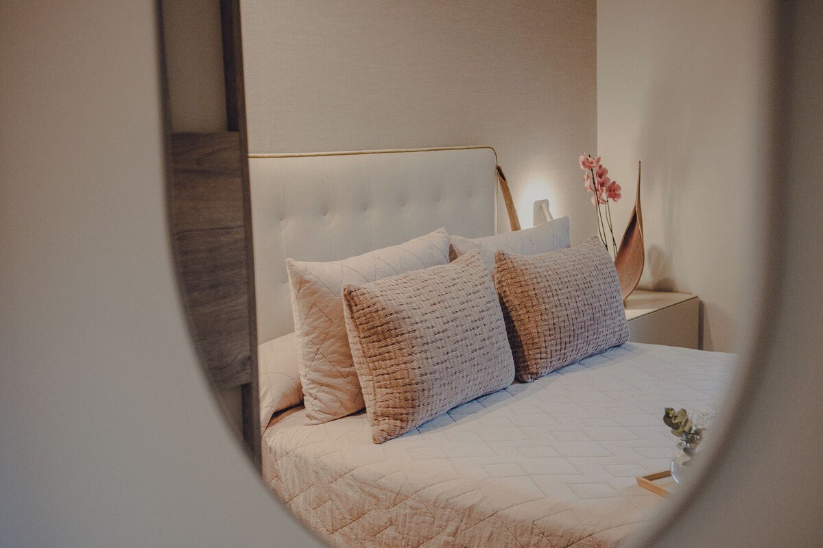 Ein ordentlich bezogenes Bett mit einer gesteppten Decke und zwei Kissen, ein Nachttisch mit Blumen in einer Vase sowie ein Spiegel, der einen Teil des Raumes reflektiert.