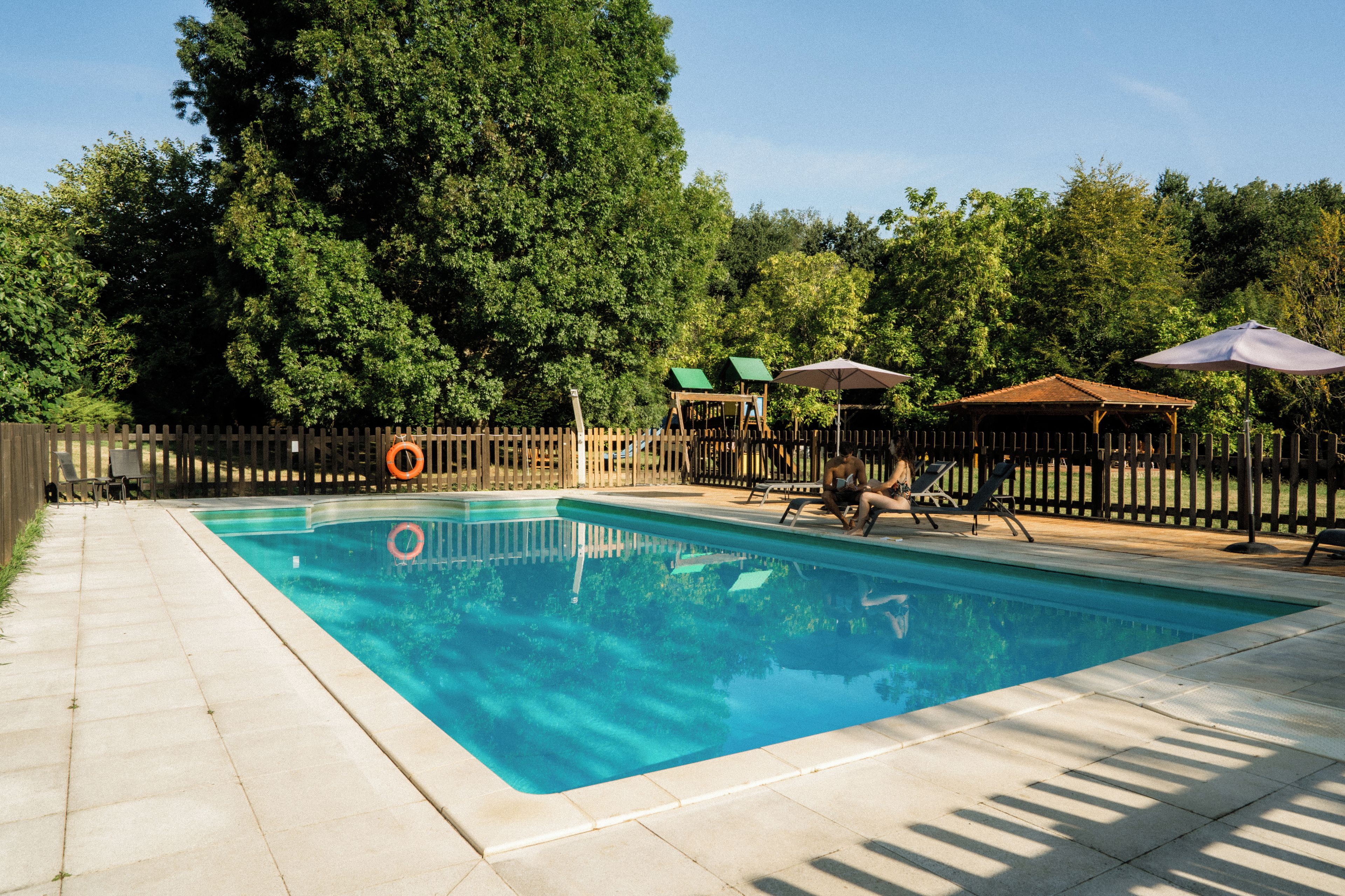 A rectangular swimming pool with clear blue water, surrounded by a wooden fence, and shaded by umbrellas.