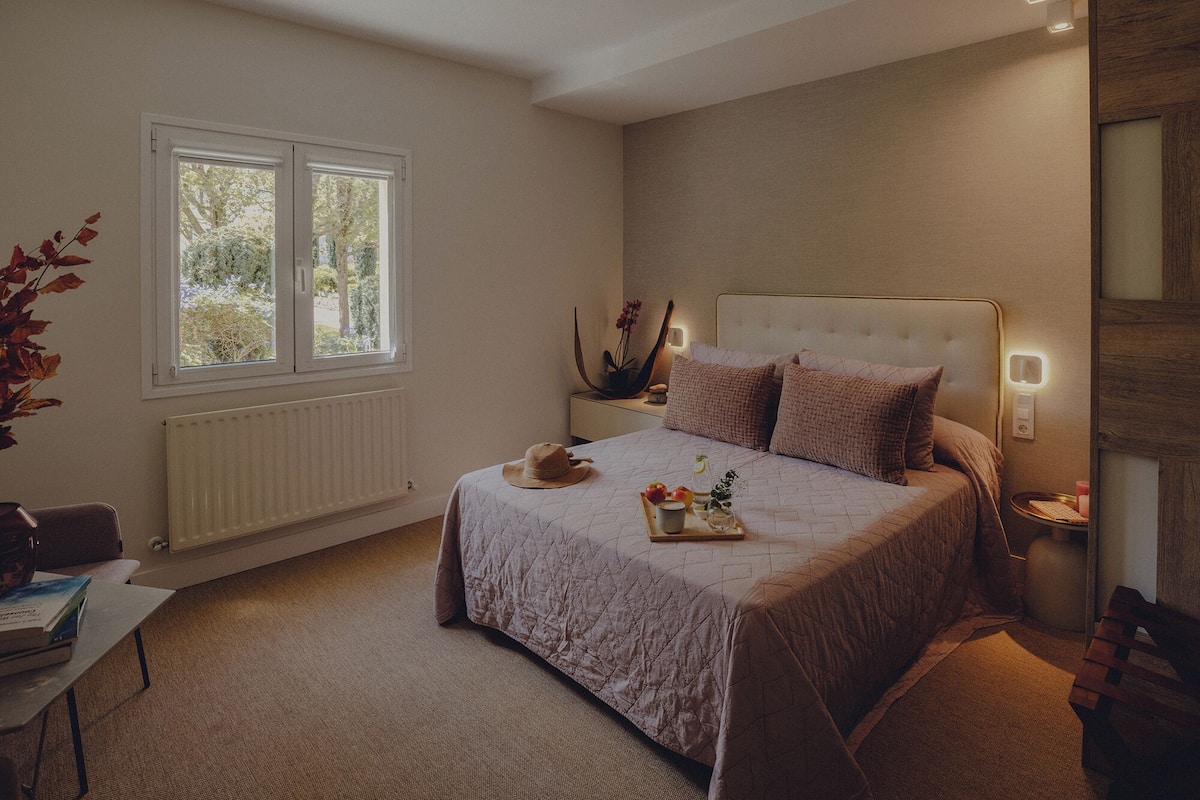 Ein Schlafzimmer mit einem großen Bett, Nachttischen, einem Heizkörper, einem Fenster und einem Schreibtisch mit Büchern.