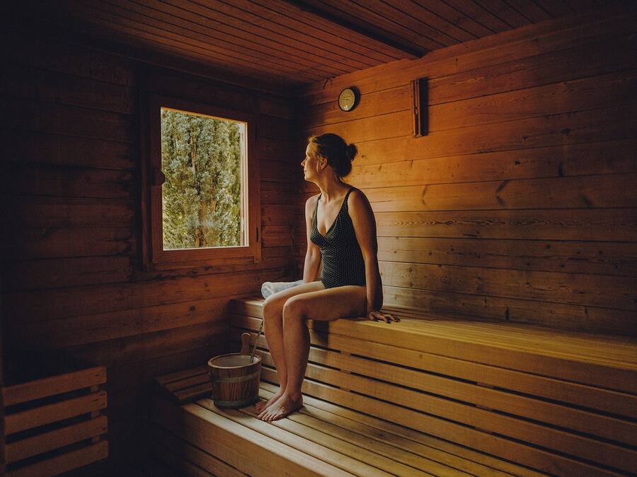 A wooden cabin with a sauna bench, a window with a view of trees, and a wooden wall clock.