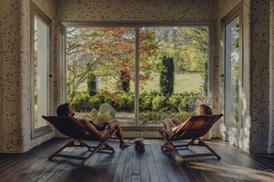 Dos personas descansando en sillones de madera junto a un ventanal con vistas a un jardín.