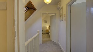 Un couloir étroit doté d’un sol recouvert de moquette, de murs blancs et d’une porte sur le côté droit. Un luminaire est fixé au plafond et une petite fenêtre se trouve sur le côté gauche.