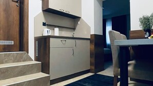 Una cocina compacta con horno empotrado, microondas y cafetera sobre la encimera. Hay una zona de comedor con mesa y sillas, y un reloj en la pared.