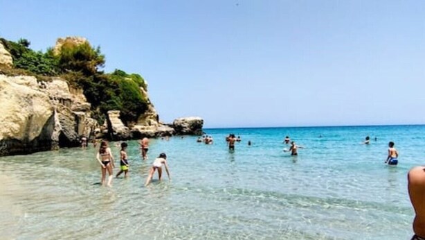 Una playa con aguas poco profundas y transparentes, acantilados rocosos y gente nadando y caminando.