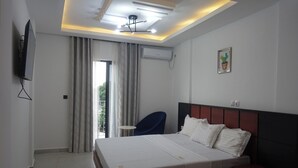 A hotel room with a bed, a chair, a TV, an air conditioner, and a window with curtains. - SMART HOTEL (Douala)