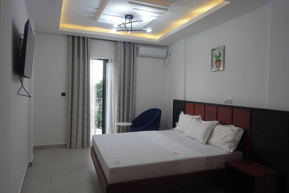 A hotel room with a bed, a chair, a TV, an air conditioner, and a window with curtains.
