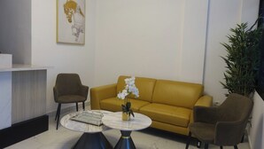 A modern lobby with a yellow sofa, two armchairs, a marble coffee table with a vase of white orchids, a framed artwork on the wall, and a potted plant. - SMART HOTEL (Douala)