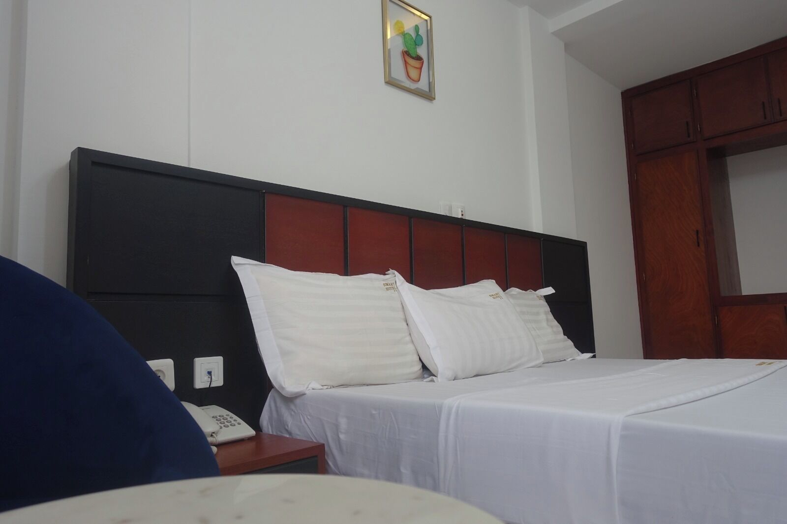 Une chambre d’hôtel avec un lit, du linge de lit blanc, une tête de lit en bois, une table de chevet avec un téléphone et un tableau accroché au mur.