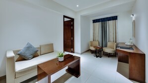 A modern living room with a sofa, coffee table, and two chairs. There is a wooden desk with a lamp and a window with curtains. - AM Hotel Kollection: Manasa Inn, Tirupati (Tirupati)