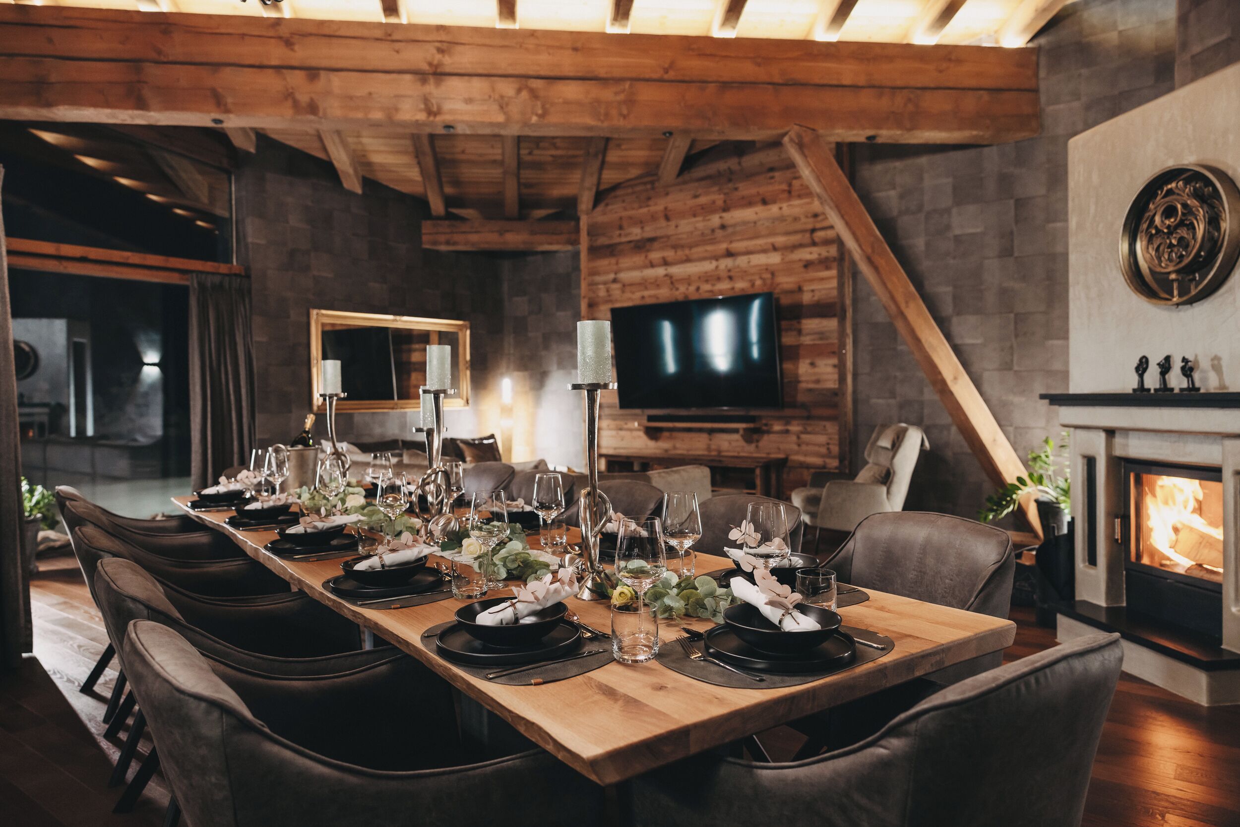 Un comedor con una larga mesa de madera dispuesta para una comida, rodeada de sillones de cuero, con una chimenea y un televisor al fondo.