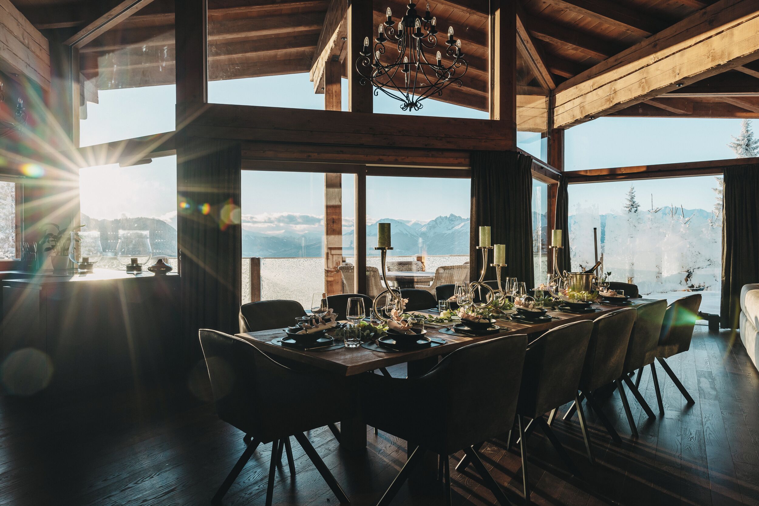 Comedor con una gran mesa de madera dispuesta para seis personas, ventanas amplias con vista a la montaña y una araña.