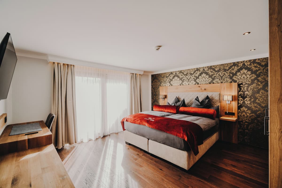 A modern hotel room with a large bed, a wooden desk, a flat-screen TV, and a large window with curtains.