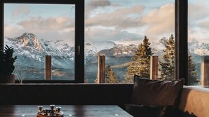 Een kamer met een groot raam met uitzicht op besneeuwde bergen, een houten tafel met kandelaar, en een gezellige zithoek.