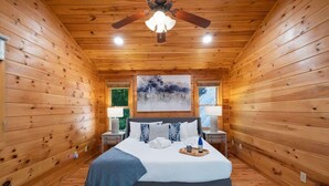 A wooden cabin bedroom with a bed, nightstands, a ceiling fan, and a painting on the wall. - Tranquil Ridge- View, pet friendly, hot tub, sauna (Epworth)