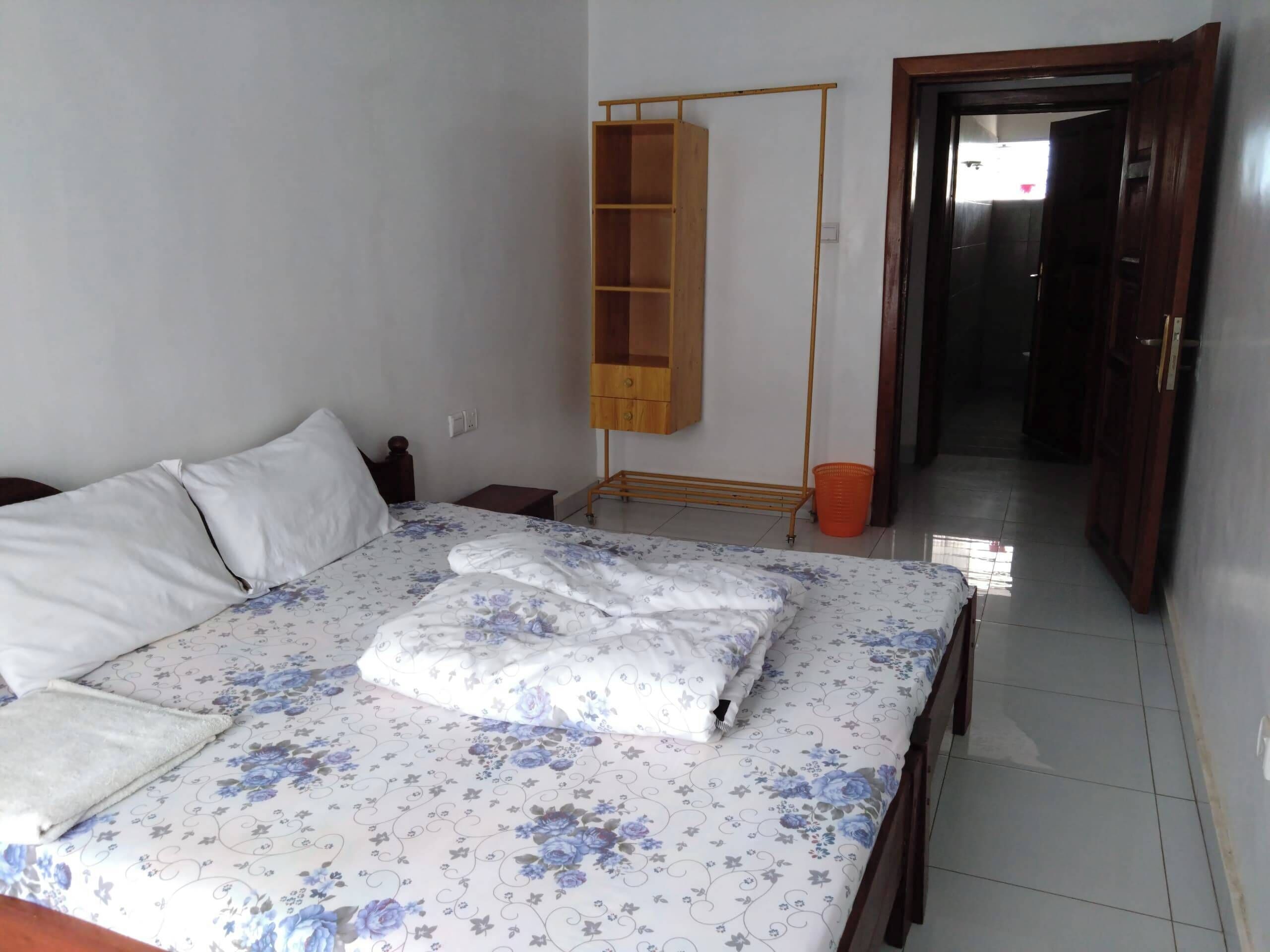 A bedroom with a bed, a wooden wardrobe, a small table, and a door leading to another room.