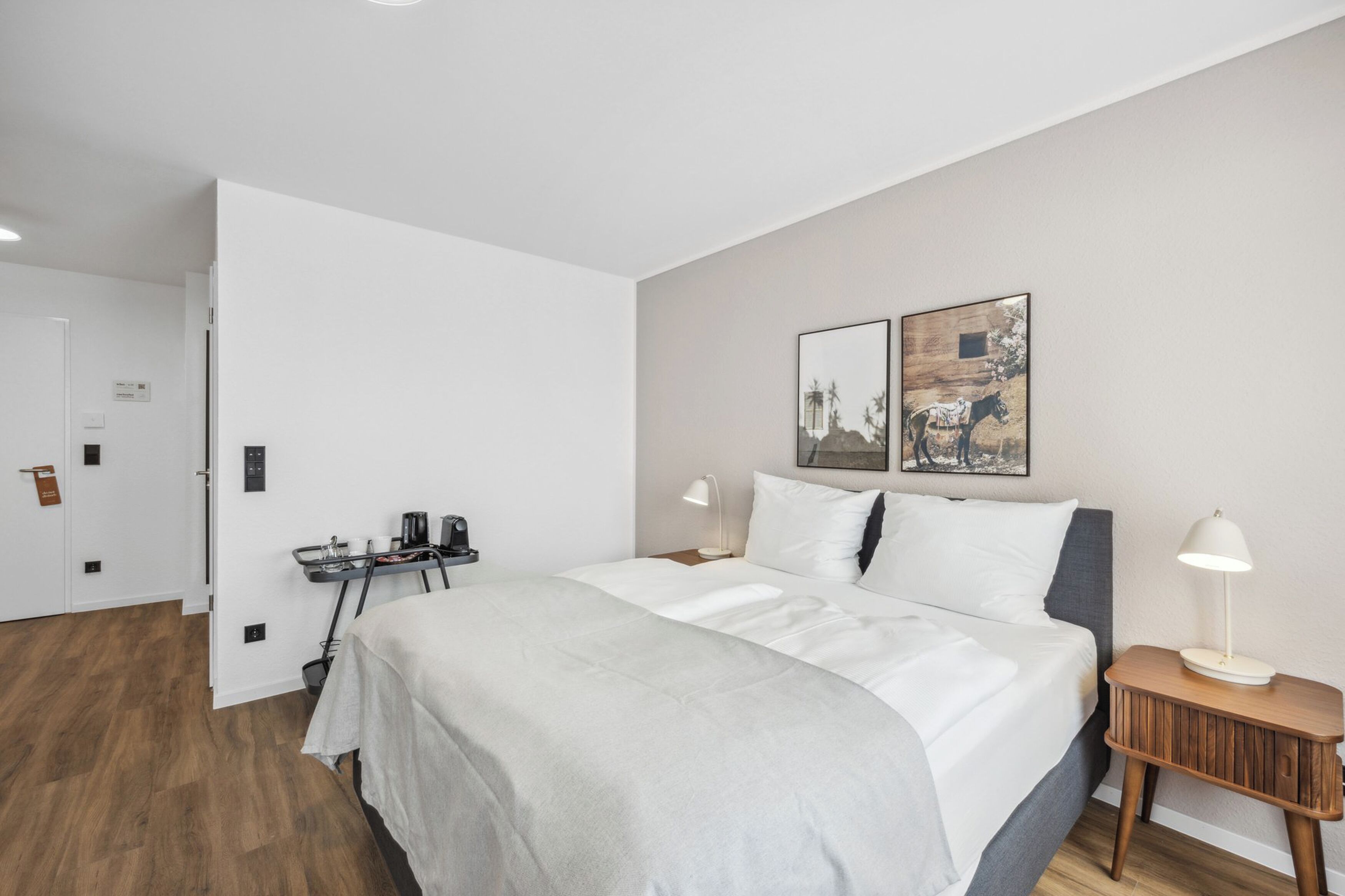 A modern hotel room with a large bed, bedside tables, a desk with a chair, and a door leading to another room.