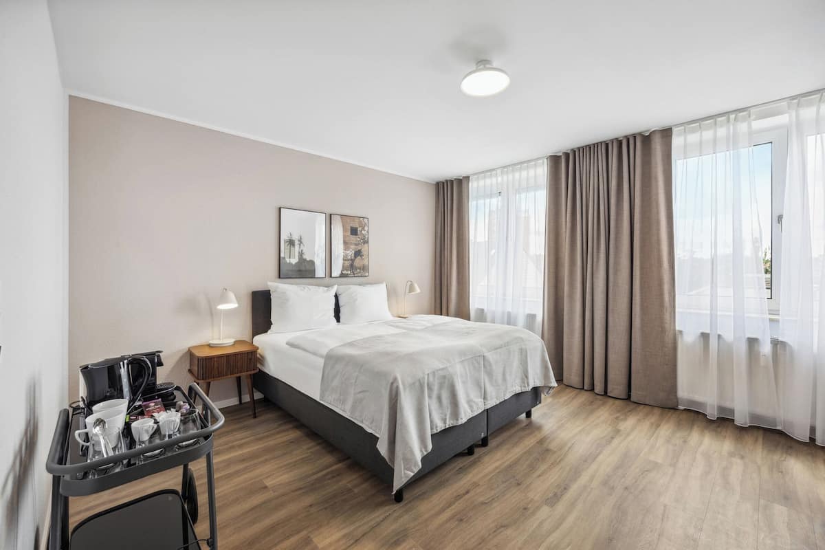 Une chambre d’hôtel moderne dotée d’un grand lit, d’une table de chevet avec une lampe, d’un petit bureau avec une machine à café et d’une fenêtre avec des rideaux.