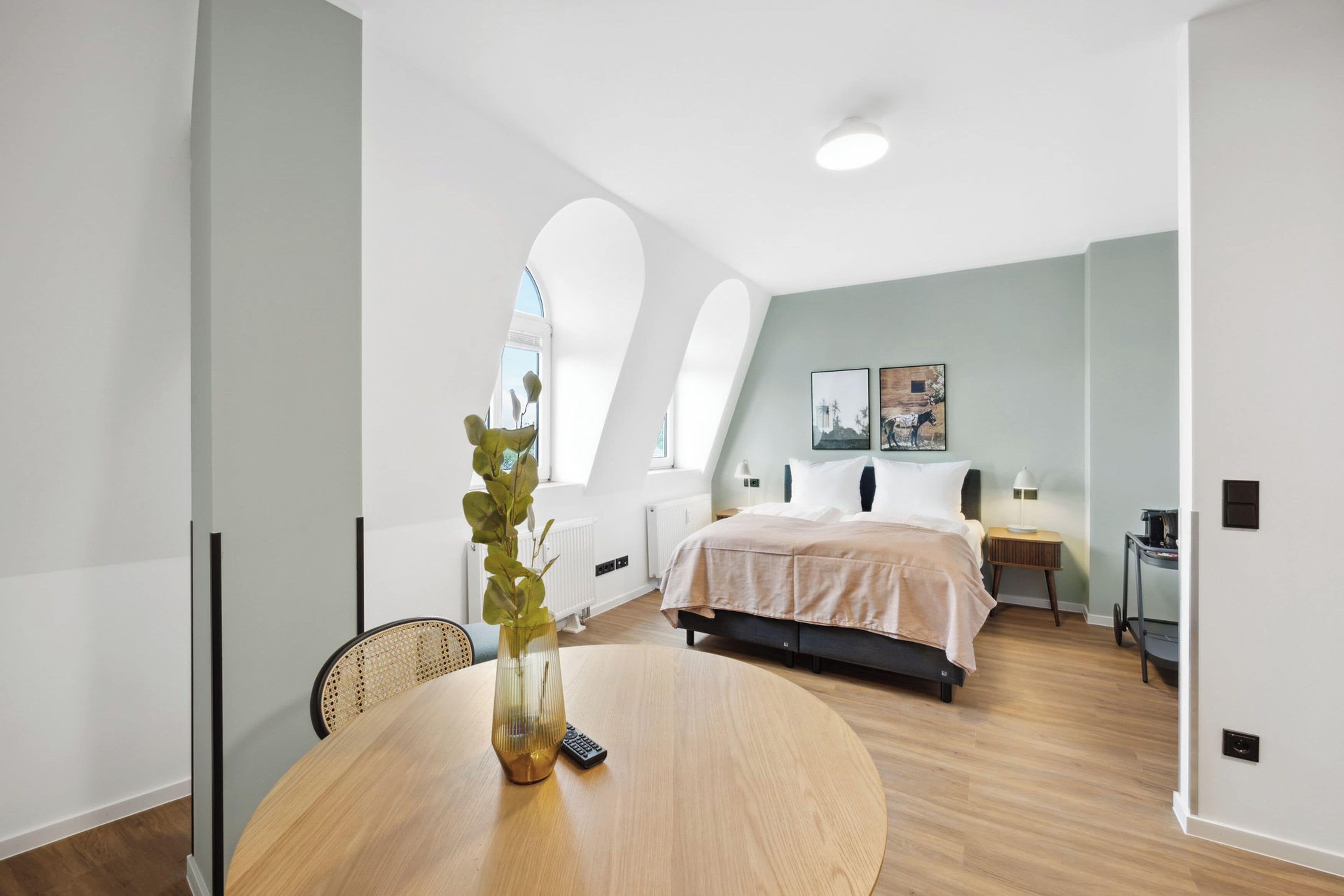 A modern bedroom with a large bed, a round wooden table with a vase and remote, and a window with a view.