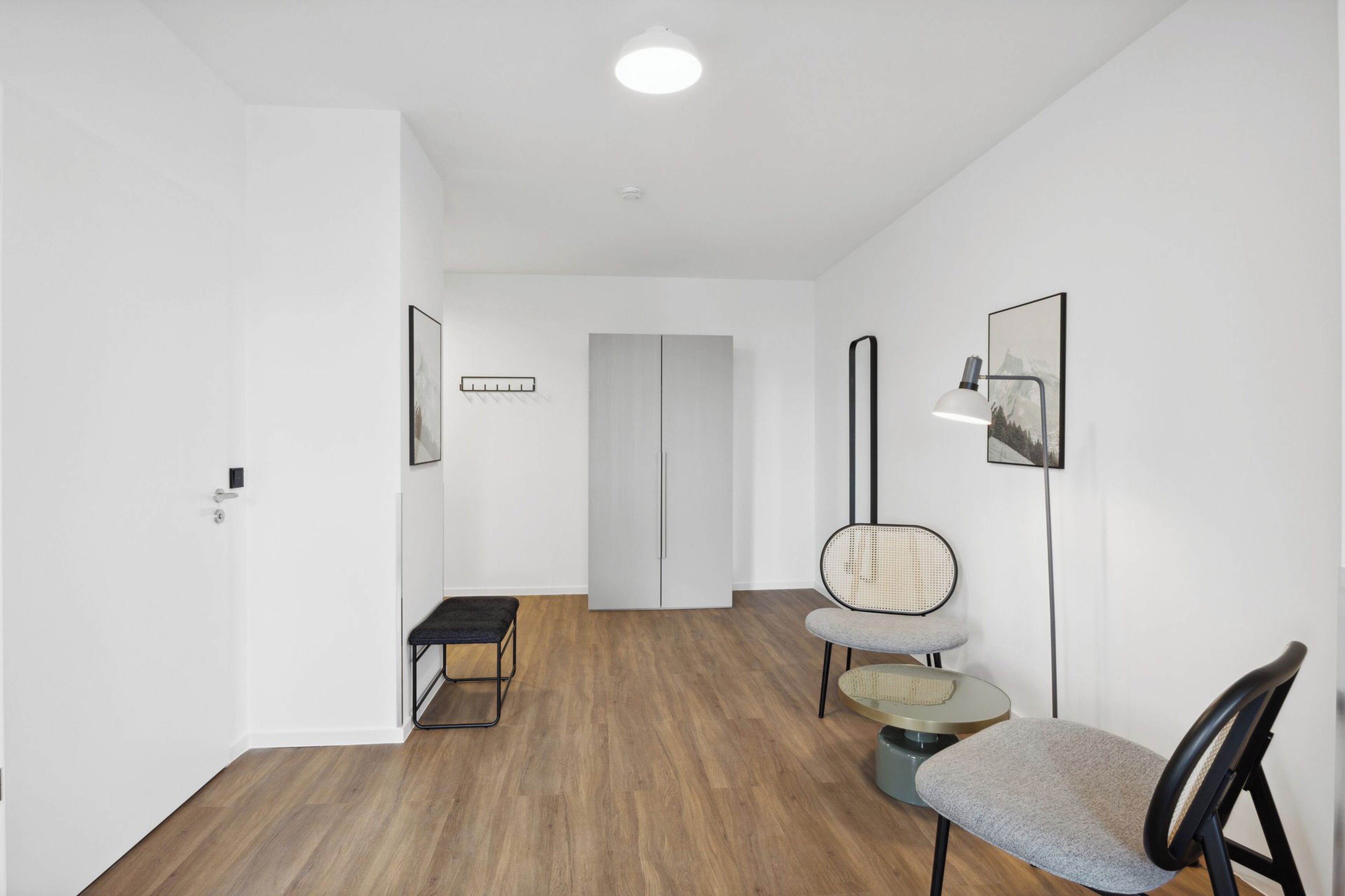 Een moderne, minimalistische gang met een houten vloer, een bankje, een ronde tafel met een stoel, en een deur aan het einde.