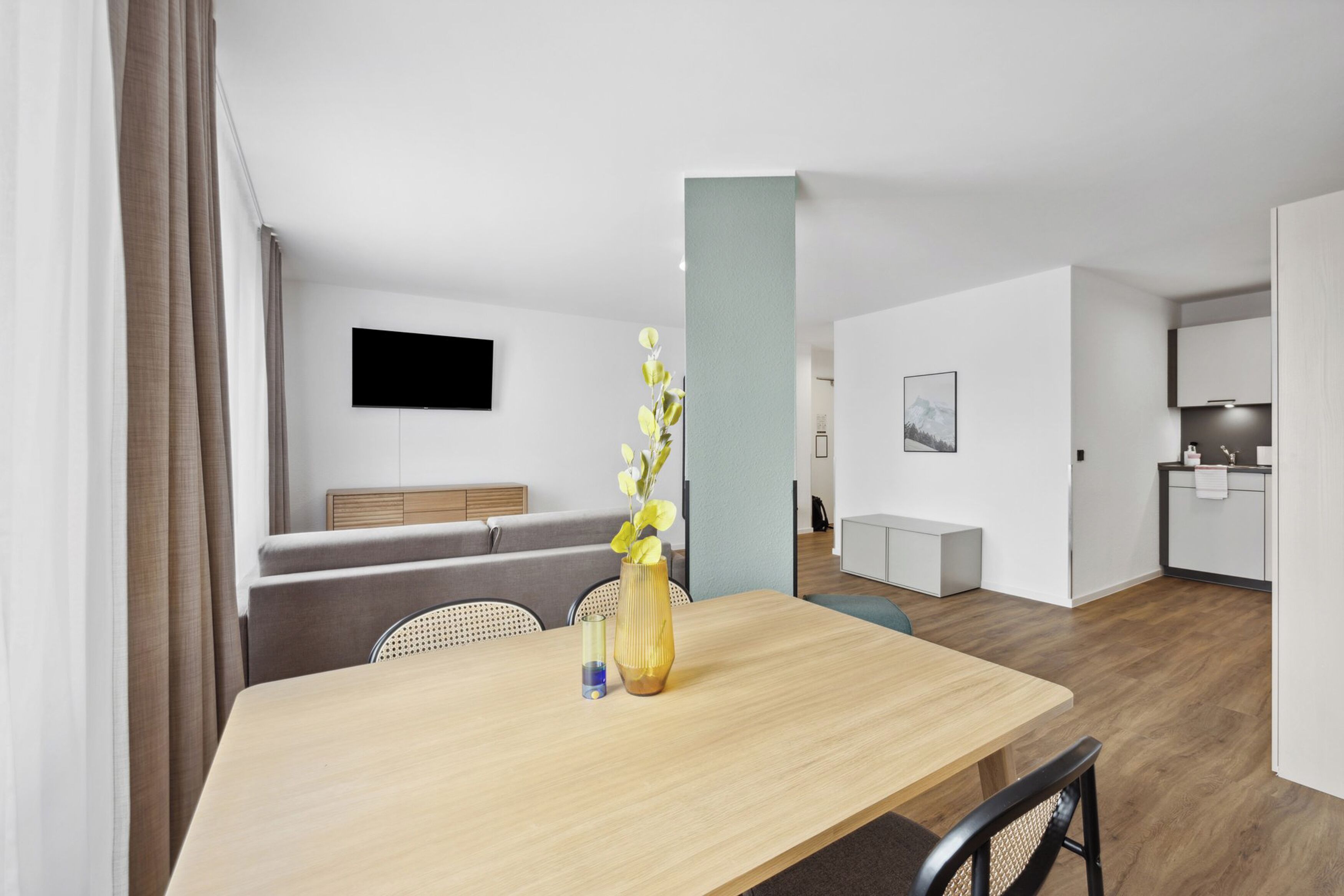 Ein modernes, helles Zimmer mit einem Holzesstisch, Stühlen, einer Blumenvase, einem an der Wand montierten Flachbildfernseher, einer Couch und einer Küchenzeile.