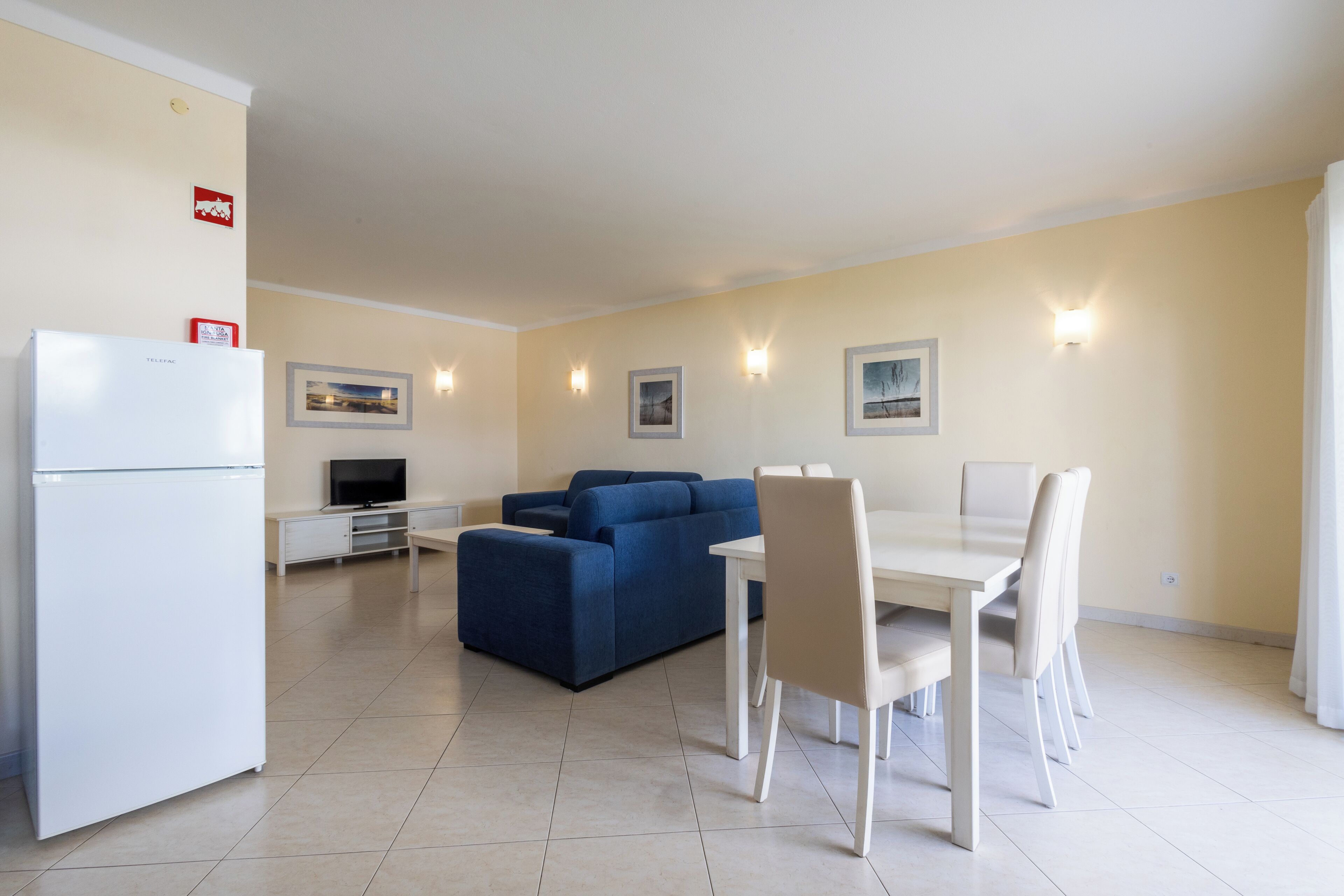 A modern living room with a blue sofa, a dining table with chairs, a white refrigerator, and wall-mounted lights.