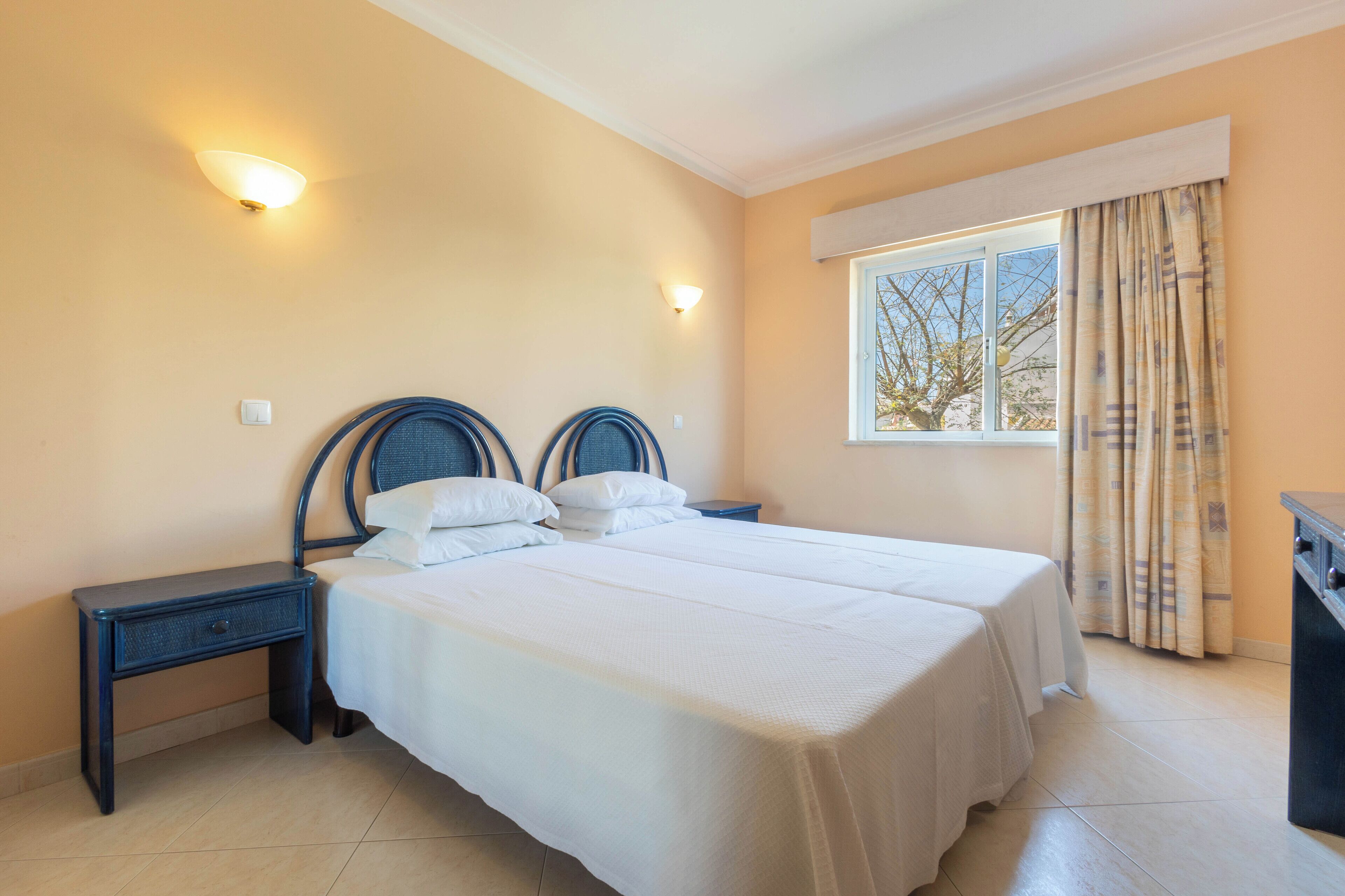 Uma cama de casal com um espalha branco, duas mesas de cabeceira e uma janela com cortinas.