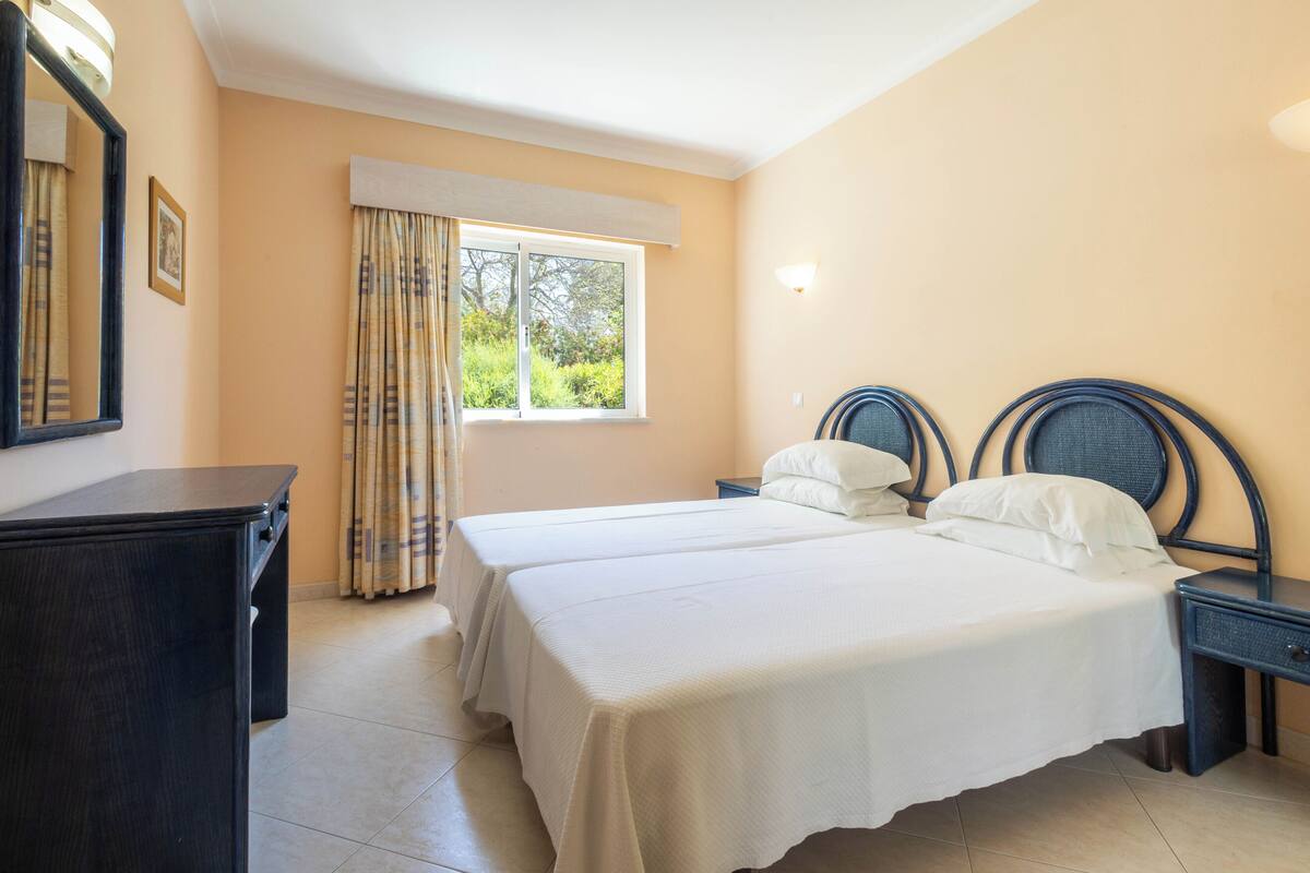 A bedroom with two beds, a window with curtains, a bedside table, and a dresser.