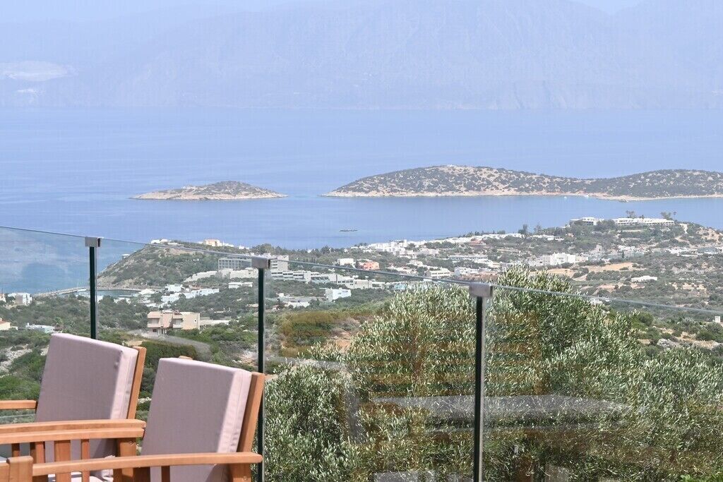 Eine Terrasse mit Stühlen, die auf eine Küstenlandschaft mit Gebäuden und Hügeln blickt.