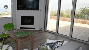 Un salon moderne avec une cheminée, un canapé agrémenté d’un coussin à motifs de feuilles, une table basse en bois et une grande fenêtre offrant une vue sur la ville.