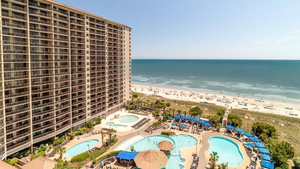 Un hôtel doté de nombreux balcons donnant sur une plage et d’un espace piscine avec des chaises longues et des parasols.