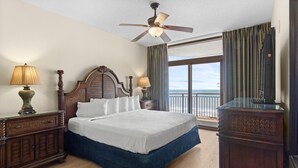 Une chambre d’hôtel avec un grand lit, une tête de lit en bois, deux lampes de chevet, un ventilateur de plafond, une grande fenêtre avec des rideaux et une vue sur l’océan.