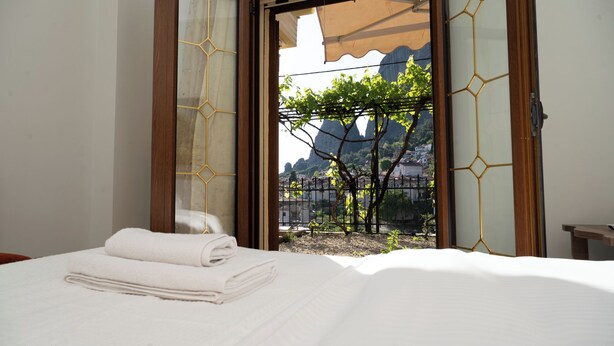 A hotel room with a bed, a window with a view of a vineyard and mountains, and a wooden door.