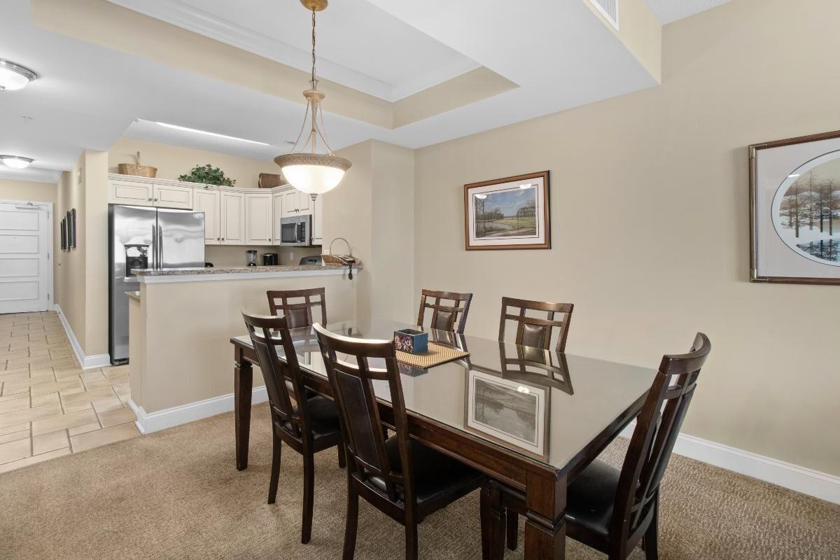A dining area with a glass table surrounded by chairs, a kitchen in the background, and a framed picture on the wall.