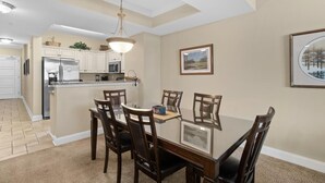 Un coin repas avec une table en verre entourée de chaises, une cuisine en arrière-plan et un tableau encadré au mur.