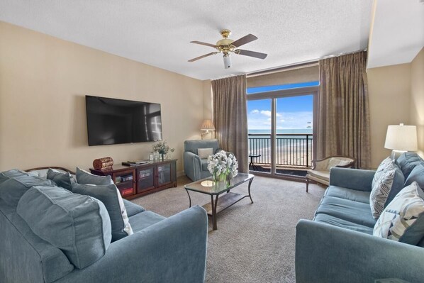 A living room with a flat-screen TV, two sofas, a coffee table, a ceiling fan, and a view of the ocean through a balcony.