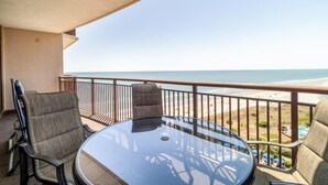 Un balcon avec une table et des chaises en verre, donnant sur une plage et l’océan.