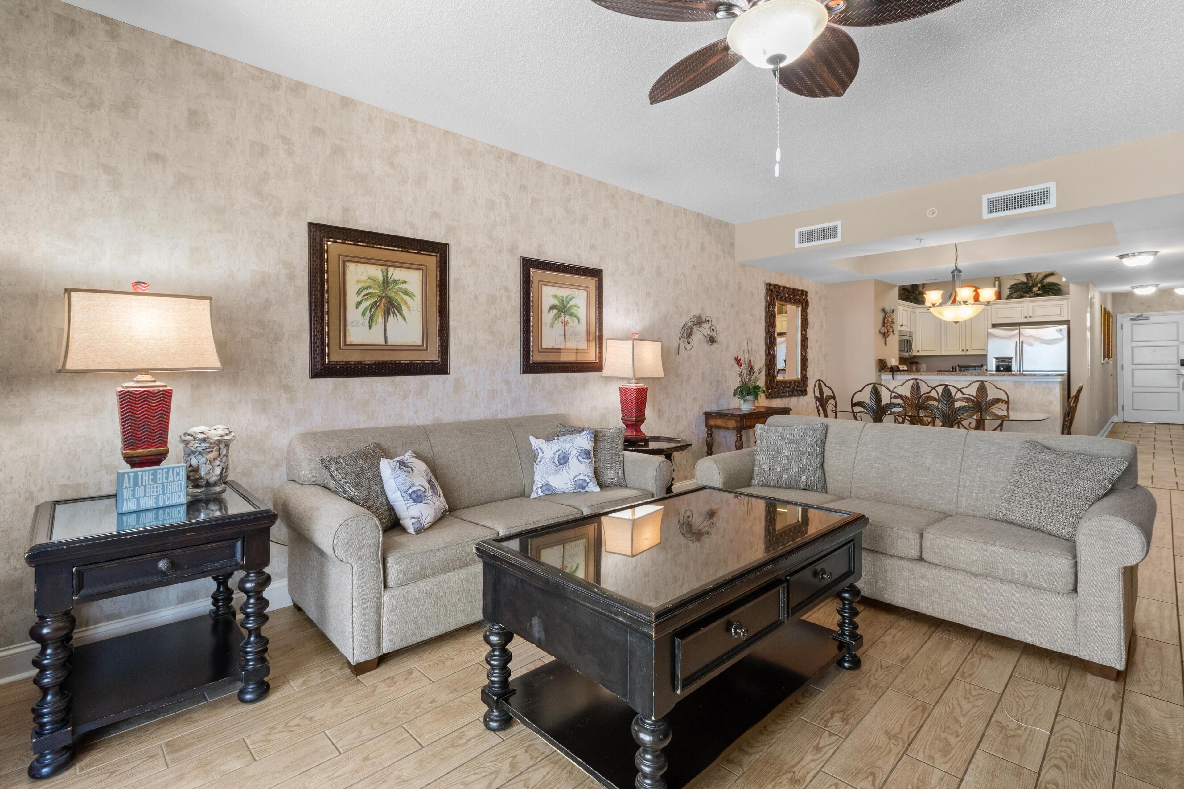 A living room with a sofa, coffee table, and two side tables with lamps. There is a ceiling fan and a kitchen in the background.