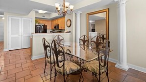 Un comedor con una mesa de vidrio rodeada de sillas ornamentales, una cocina al fondo y un espejo grande en la pared.