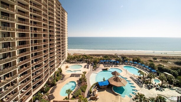 Un hotel de gran altura con múltiples balcones que dan a una playa y un área de piscina con diversas fuentes.
